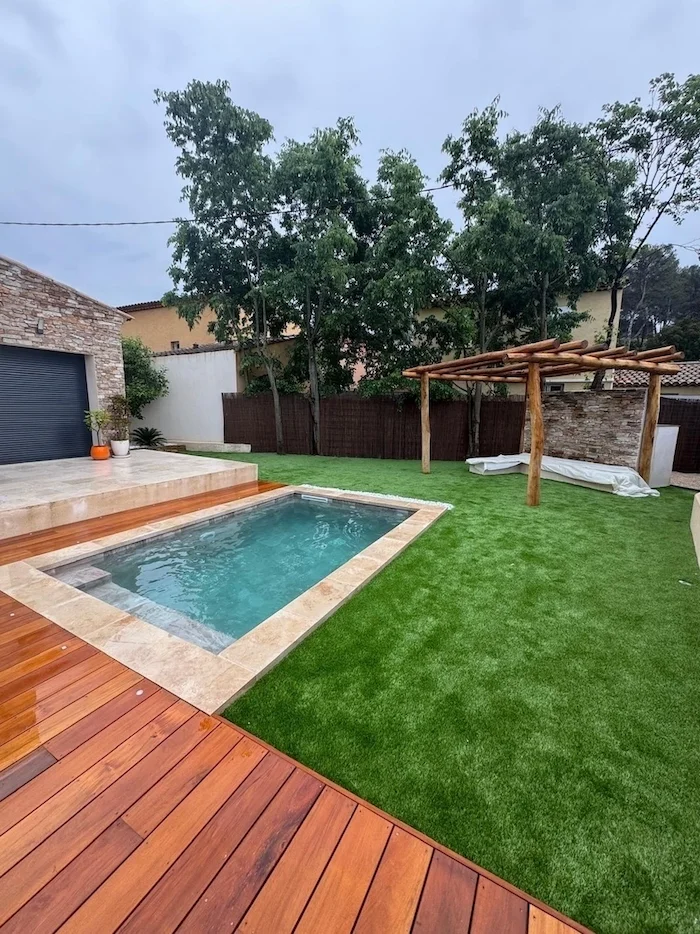 Petite piscine avec terrasse en bois, pelouse et pergola dans un jardin clos.